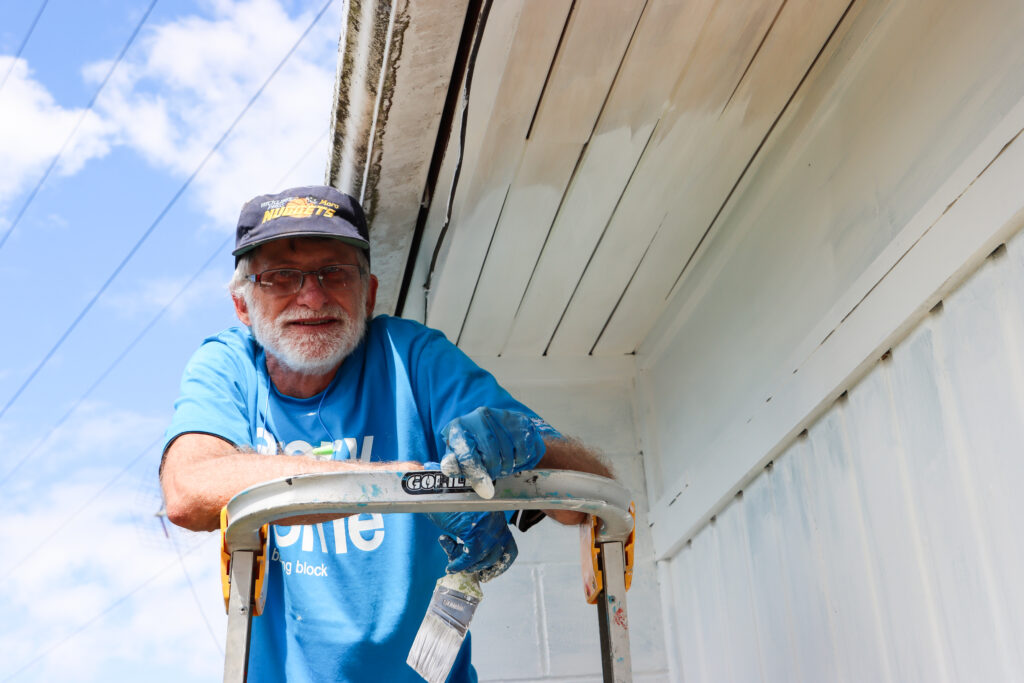 Volunteer smiles at the camera as he paints the exterior of a home. 